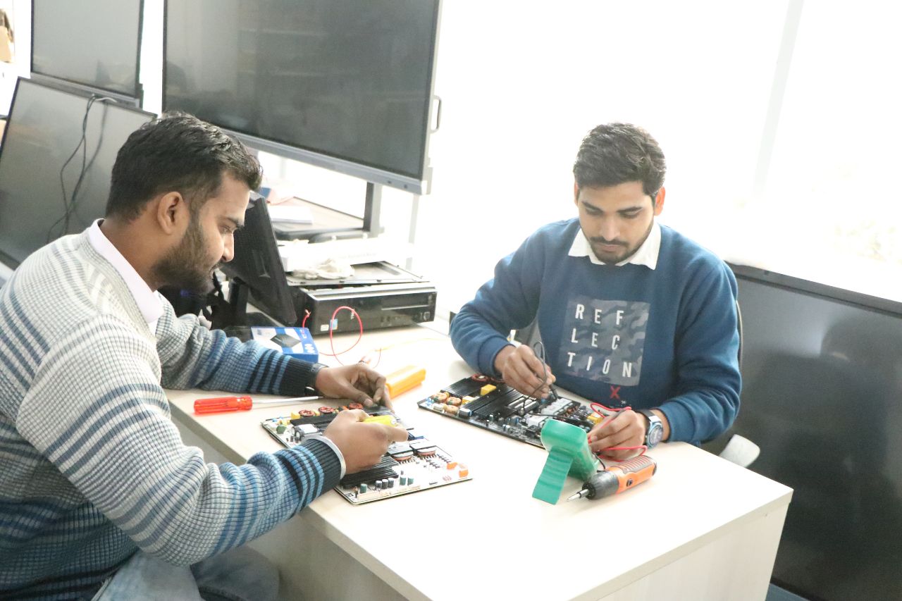 Two engineers testing circuit boards at a workbench beside interactive panels